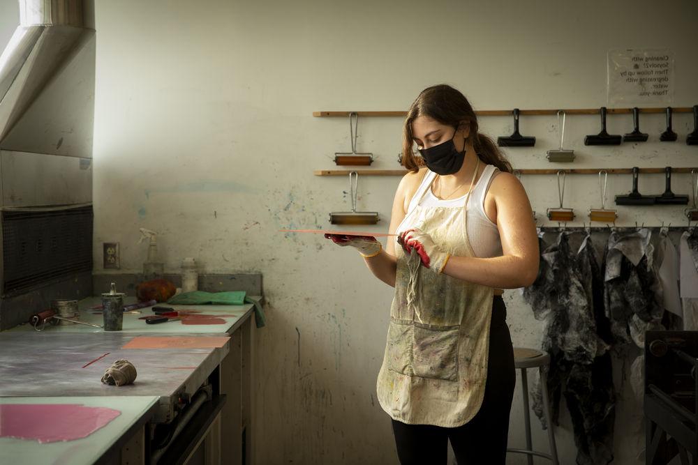 一名戴着口罩和手套的学生在美术工作室拿着一个方形的印版.